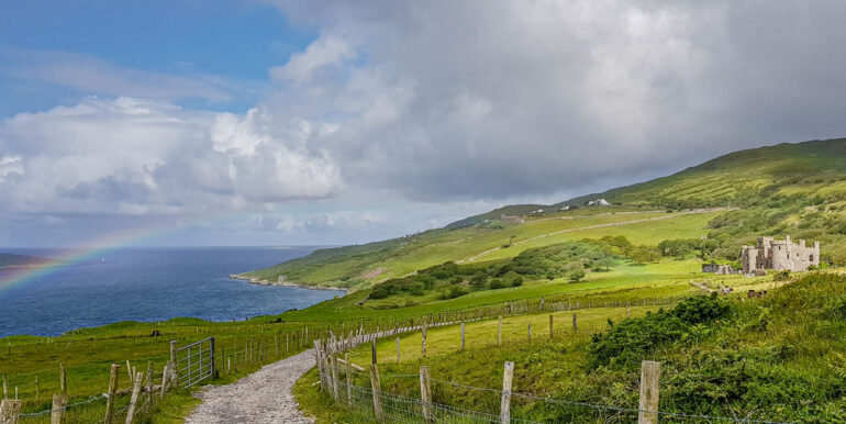Stone pathway with wooden pole fence and barbed wire between the Irish countryside and the sunlit Clifden Castle in the background